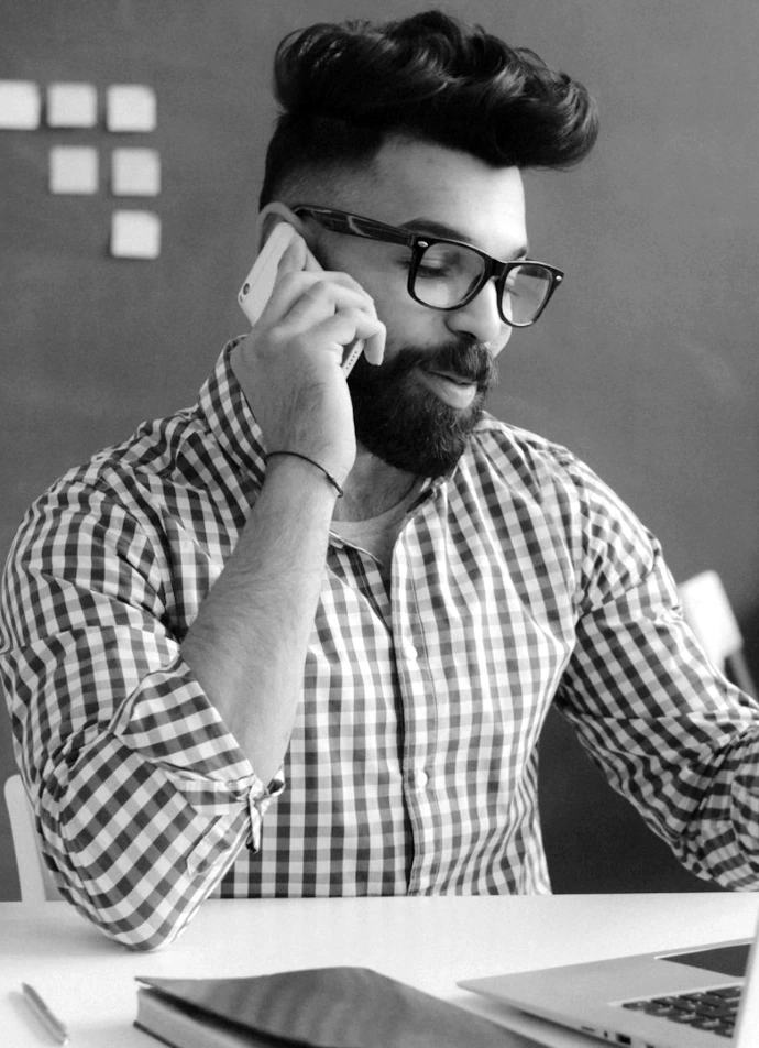 A man with a beard and glasses talking on a phone at a desk